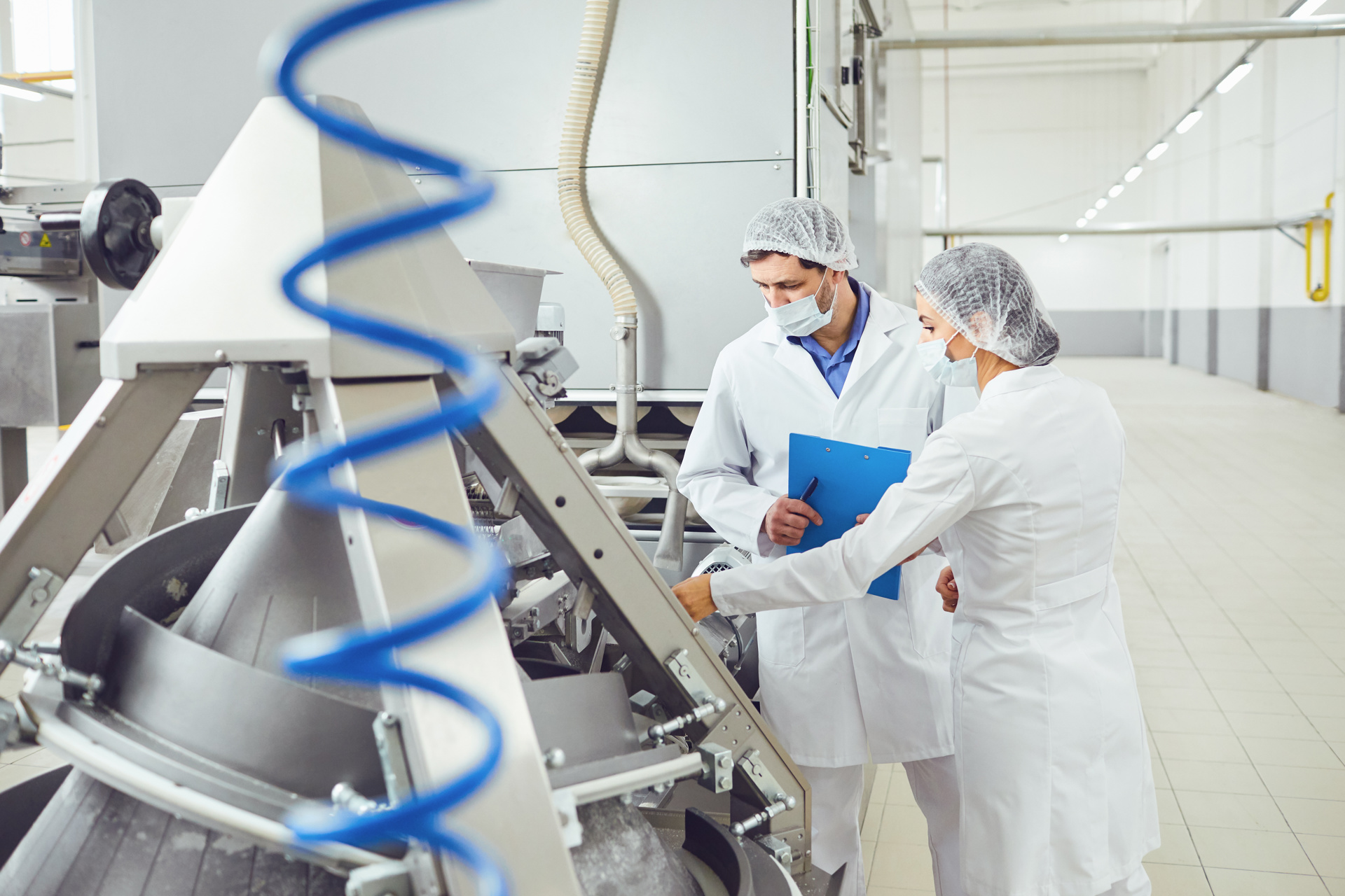 Zwei Mitarbeiter in Schutzkleidung und Haarnetzen besprechen sich vor einer großen industriellen Maschine in einer Produktionshalle