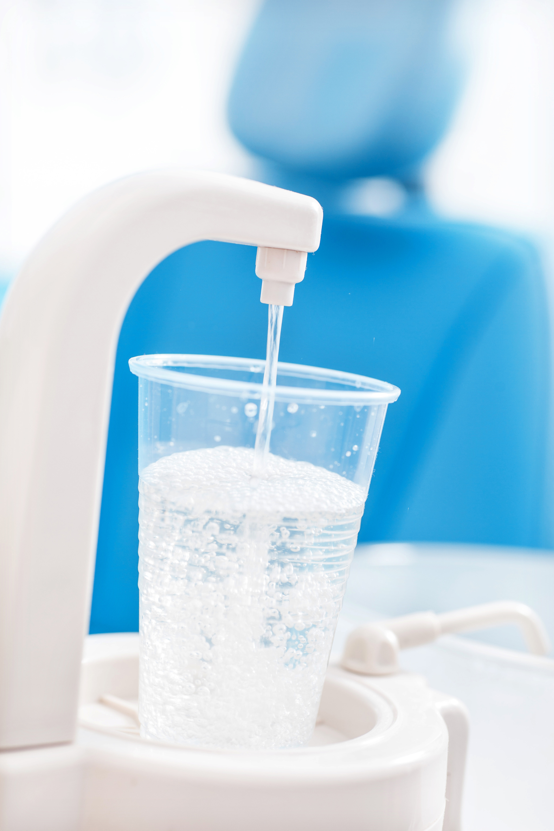 Wasser läuft aus einem Wasserhahn in einen durchsichtigen Becher vor einem blauen Zahnarztstuhl
