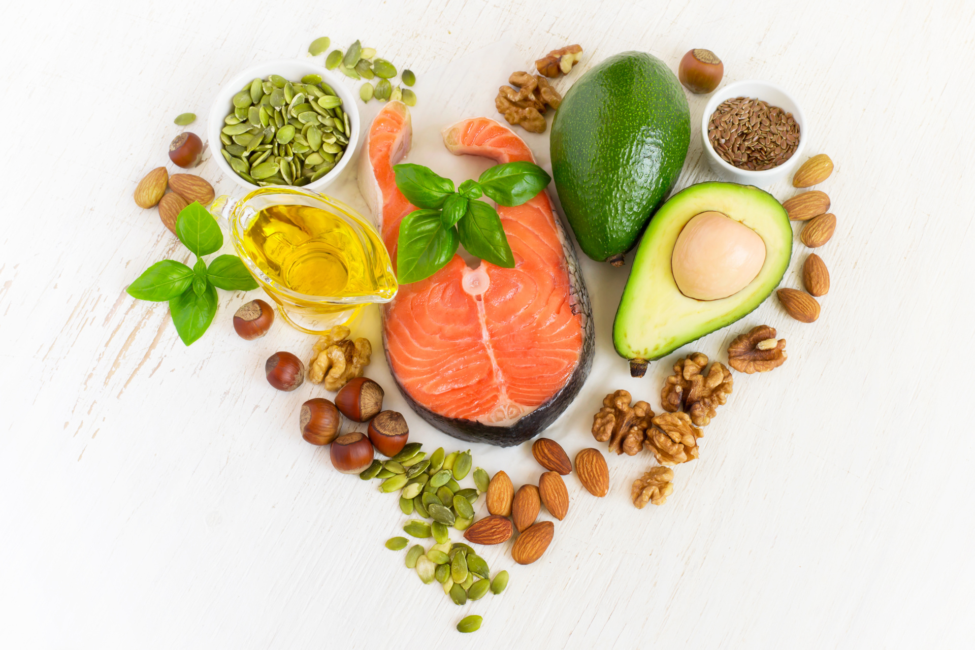 Herzförmige Anordnung von gesunden Lebensmitteln wie Lachsfilet, Avocado, Nüssen, Samen und Olivenöl auf weißem Tisch